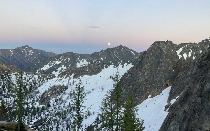 A full moon appears above snowy mountains in a sky illuminated in soft blues and purples. 